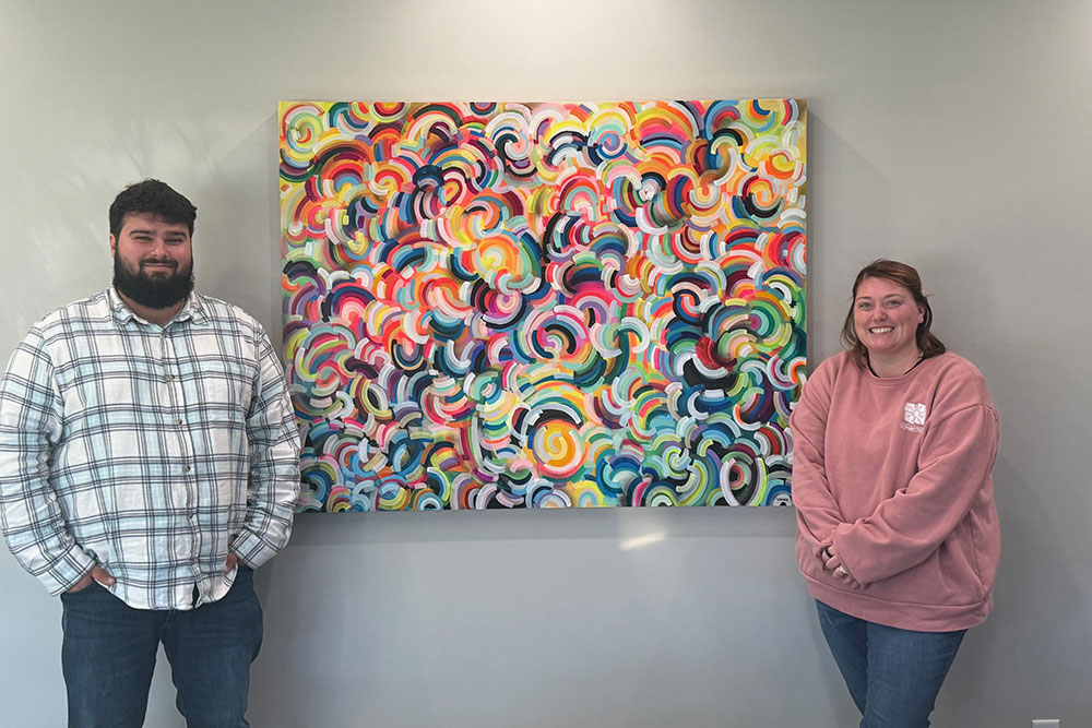 Two coworkers stand on either side of a large, colorful abstract painting filled with overlapping rainbow-like swirls. The man on the left wears a plaid button-down shirt and jeans, and the woman on the right wears a pink sweatshirt and jeans. They are smiling and standing against a light gray wall.