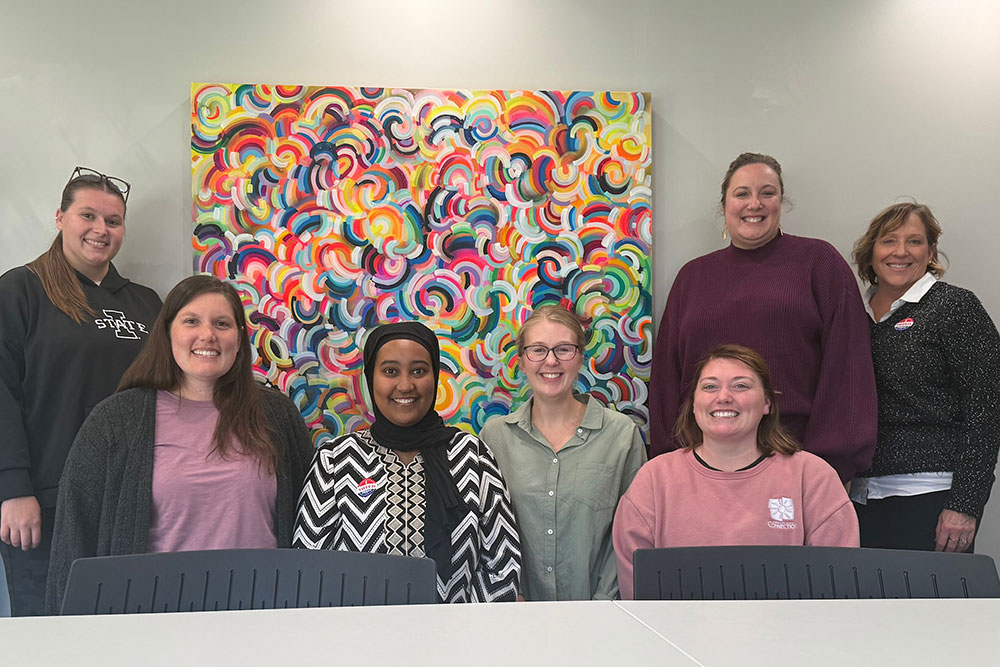 Seven coworkers pose together in front of a large, vibrant abstract painting made up of layered, multicolored swirl patterns. Four women stand in the back row and three sit in front behind a white table. Everyone is smiling, and the group is positioned against a light gray wall.